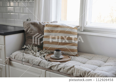 Moody epring interior still life. Cup of coffee, tea on wooden tray. Blossoming cherry plum tree branches. Sitting bench with linen cushions under window in kitchen. Easter breakfast, home decor. Top Moody epring interior still life. Cup of coffee, tea on wooden tray. Blossoming cherry plum tree branches. Sitting bench with linen cushions under window in kitchen. Easter breakfast, home decor. Top 112926458