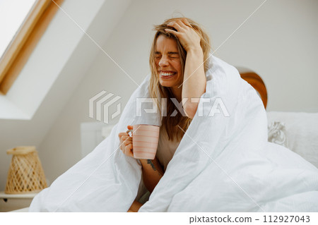 Woman in formal wear smiles, sitting on white bedding with coffee cup Woman in formal wear smiles, sitting on white bedding with coffee cup 112927043