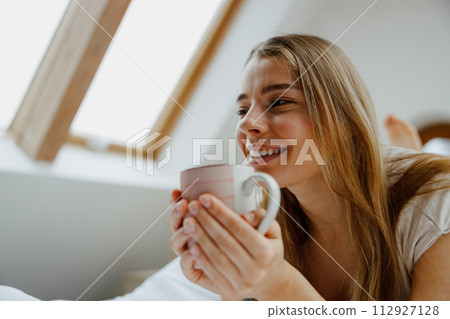 Young woman is sitting on a bed holding a cup of coffee Young woman is sitting on a bed holding a cup of coffee 112927128