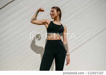 Smiling female model in sportswear showing biceps standing outdoors and looks away 112927187