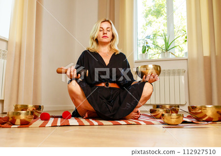 Woman playing on a tibetian singing bowl in cozy room meditating in a yoga Woman playing on a tibetian singing bowl in cozy room meditating in a yoga 112927510