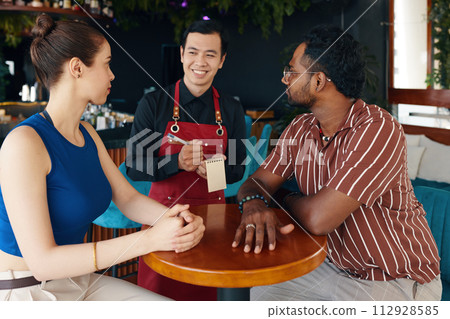 Smiling waiter talking to couple sitting at restaurant table 112928585