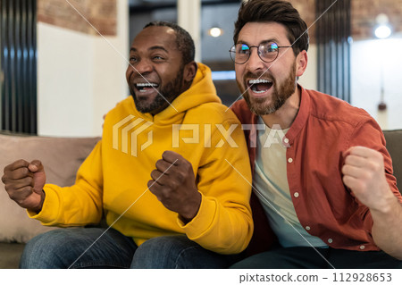 Two friends sitting together and looking excited while watching tv Two friends sitting together and looking excited while watching tv 112928653