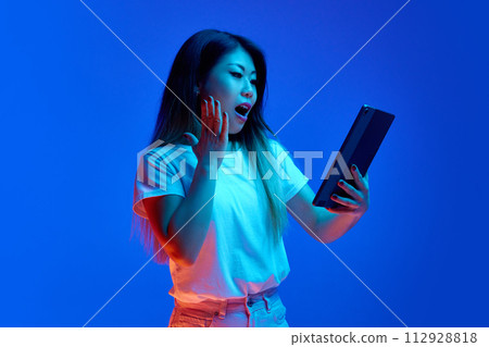 Young Asian woman looking to tablet with shocked facial expression against blue gradient background in neon light. Young Asian woman looking to tablet with shocked facial expression against blue gradient background in neon light. 112928818