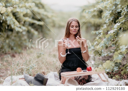 Woman picnic vineyard. Happy woman with a glass of wine at a picnic in the vineyard, wine tasting at sunset and open nature in the summer. Romantic dinner, fruit and wine. 112929180