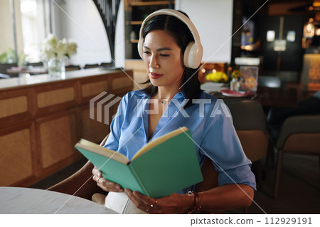 Woman sitting at restaurant table listening to calm music in headphones and reading planner Woman sitting at restaurant table listening to calm music in headphones and reading planner 112929191