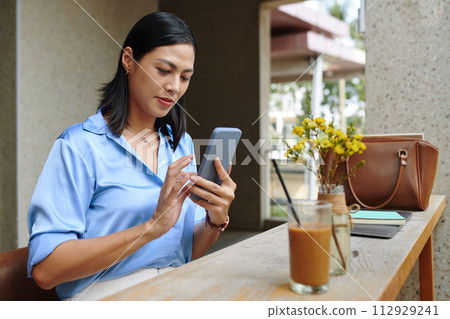 Businesswoman sitting at outdoor cafe table and checking messages in smartphone 112929241