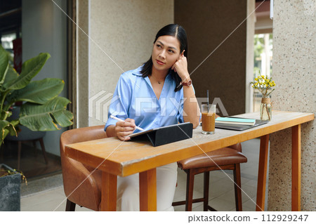 Pensive woman sitting at cafe table and sketching logo of her new company on tablet 112929247
