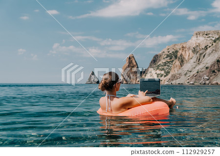 Woman freelancer works on laptop swimming in sea on pink inflatable ring. Happy tourist in sunglasses floating on inflatable donut and working on laptop computer in calm ocean. Remote working anywhere 112929257