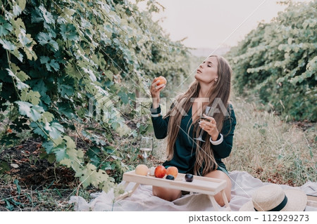Woman picnic vineyard. Happy woman with a glass of wine at a picnic in the vineyard, wine tasting at sunset and open nature in the summer. Romantic dinner, fruit and wine. Woman picnic vineyard. Happy woman with a glass of wine at a picnic in the vineyard, wine tasting at sunset and open nature in the summer. Romantic dinner, fruit and wine. 112929273