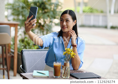 Smiling Vietnamese businesswoman taking selfie at outdoor cafe table 112929274