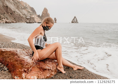 Woman travel portrait. Happy woman with long hair poses on a red volcanic rock at the beach. Close up portrait cute woman in black bikini, smiles at the camera, with the sea in the background. Woman travel portrait. Happy woman with long hair poses on a red volcanic rock at the beach. Close up portrait cute woman in black bikini, smiles at the camera, with the sea in the background. 112929275