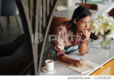 Pensive female entrepreneur listening to music and filling gratitude journal 112929285
