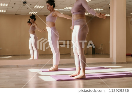 Well looking middle aged sporty woman, fitness instructor in pink sportswear doing stretching and pilates on yoga mat in the studio with mirror. Female fitness yoga. Healthy lifestyle and harmony. 112929286