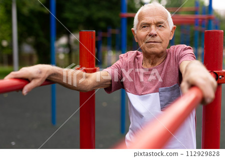Old man standing between parallel bars outdoors Old man standing between parallel bars outdoors 112929828