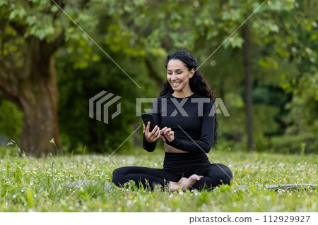 Smiling woman dressed in black activewear sitting on grass, engaging with her smartphone in a lush green park. 112929927