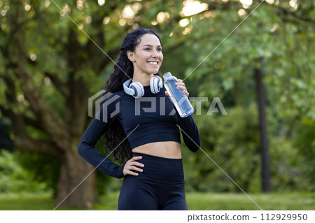 Smiling active woman in workout gear taking a break with a water bottle in a green park setting, headphones around her neck. 112929950