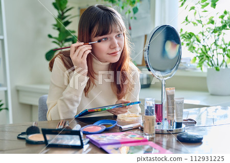 Young woman with cosmetic products, doing makeup looking in mirror at home 112931225