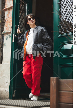 Fashionable young woman wearing stylish red cargo pants and bomber jacket, exiting door. Outdoor clothing brand showcasing versatile street-to-trail apparel. 112931600