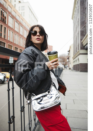 Young woman in stylish gray jacket, metal colored bag and red pants standing with coffee cup, walking around city. 112931607