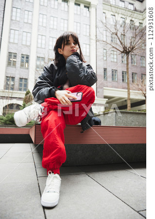Young woman in stylish gray jacket and red pants sitting on bench with mobile phone against urban background Young woman in stylish gray jacket and red pants sitting on bench with mobile phone against urban background 112931608