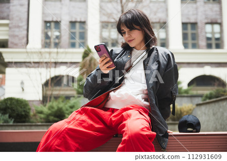 Young woman in stylish gray jacket and red pants sitting on bench with mobile phone against urban background Young woman in stylish gray jacket and red pants sitting on bench with mobile phone against urban background 112931609