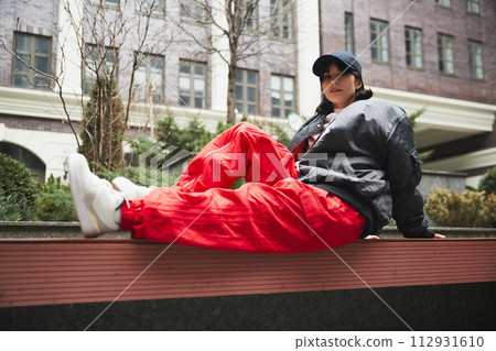 Contemporary urban clothing outlets. Stylish beautiful young woman sitting on bench in red cargo pants, stylish bomber jacket and cap 112931610