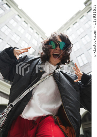 Joyful young woman in red pants and black jacket, making emotional face against sky background. Street style Joyful young woman in red pants and black jacket, making emotional face against sky background. Street style 112931615