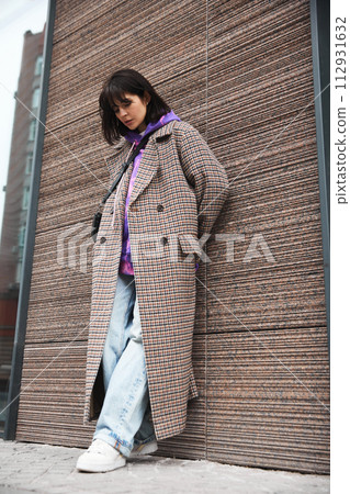Young brunette woman with sort hair wearing stylish plaid coat, hoodie and jeans, posing against textured street wall Young brunette woman with sort hair wearing stylish plaid coat, hoodie and jeans, posing against textured street wall 112931632