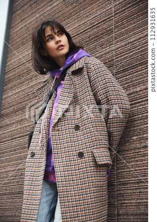 Young brunette woman with sort hair wearing stylish plaid coat, hoodie and jeans, posing against textured street wall 112931635