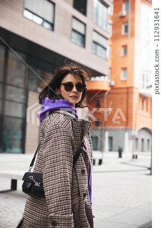 Stylish young woman in trendy sunglasses, in coat and hoodie, with stylish bag posing against urban background. Windy weather Stylish young woman in trendy sunglasses, in coat and hoodie, with stylish bag posing against urban background. Windy weather 112931641
