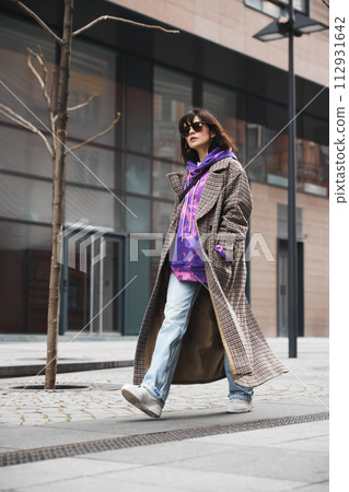 Urban fashion show. Young woman in dynamic pose wearing coat, jeans and bright hoodie walking along empty street Urban fashion show. Young woman in dynamic pose wearing coat, jeans and bright hoodie walking along empty street 112931642