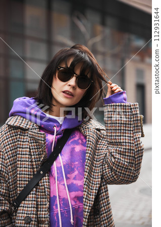 Portrait of young brunette woman in stylish sunglasses, colorful hoodie and classic coat posing against urban background Portrait of young brunette woman in stylish sunglasses, colorful hoodie and classic coat posing against urban background 112931644