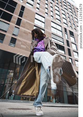Creative street photography with young woman in stylish casual clothes, coat, jeans and hoodies posing against urban architecture 112931679