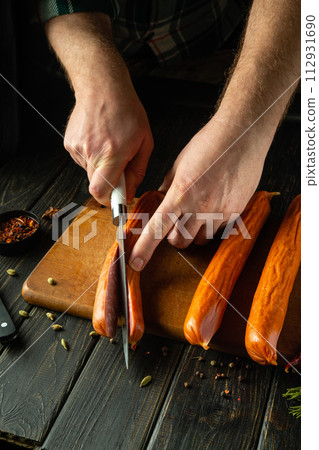Slicing sausage with a knife in the hand of a cook on a kitchen board before preparing dinner. Low key concept for serving meat dish Slicing sausage with a knife in the hand of a cook on a kitchen board before preparing dinner. Low key concept for serving meat dish 112931690
