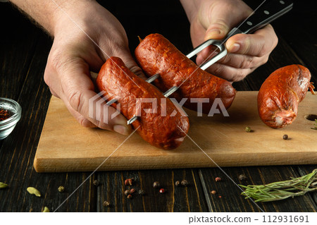 Close-up of a chef hands with meat sausages and a fork for delicious grilling. Low key barbecue cooking concept with sausages 112931691