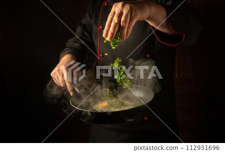 Cropped silhouette of a chef with a hot frying pan for cooking scrambled eggs for breakfast. The cook hand tosses fresh parsley into fried eggs for aroma and taste Cropped silhouette of a chef with a hot frying pan for cooking scrambled eggs for breakfast. The cook hand tosses fresh parsley into fried eggs for aroma and taste 112931696