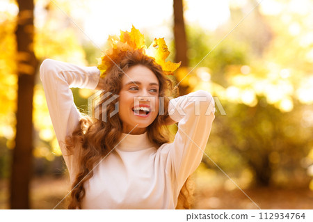 Smiling young woman with the fall crown of yellow leaves on her curly hairstyle head in the autumn park at sunset Smiling young woman with the fall crown of yellow leaves on her curly hairstyle head in the autumn park at sunset 112934764