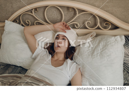 Young woman with headache flu ill sick disease cold at home indoor lying on bed with cold compress on her head at bedroom home Young woman with headache flu ill sick disease cold at home indoor lying on bed with cold compress on her head at bedroom home 112934807