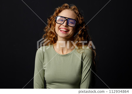 Happy smiling curly young woman wearing glasses looking at camera with joyful smile at black background Happy smiling curly young woman wearing glasses looking at camera with joyful smile at black background 112934864