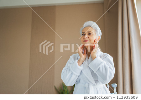 Mature adult old woman applying nourishing anti age face cream doing daily beauty routine. Senior woman touching face skin wrinkles gets facial treatment. Skin care aging beauty skincare cosmetics Mature adult old woman applying nourishing anti age face cream doing daily beauty routine. Senior woman touching face skin wrinkles gets facial treatment. Skin care aging beauty skincare cosmetics 112935609