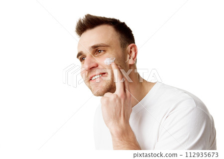 Young handsome man applying facial care, day cream and looking away against white studio background. Young handsome man applying facial care, day cream and looking away against white studio background. 112935673