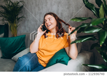 Happy young woman talk phone, listen, sit sofa indoors house. Phone Communication and comfort concept. Feminine gossip girl while relaxing on couch at home 112936740