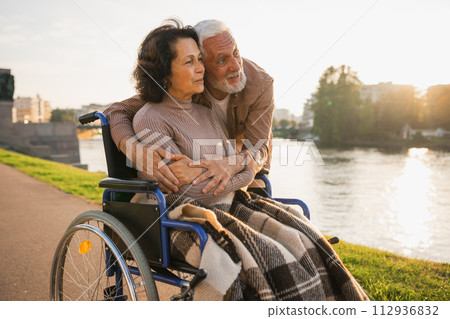 Senior woman in wheelchair walking with caregiver old man on road in park. Elderly family couple man supporting embracing woman in chair for people with disability outdoor. Rehabilitation 112936832