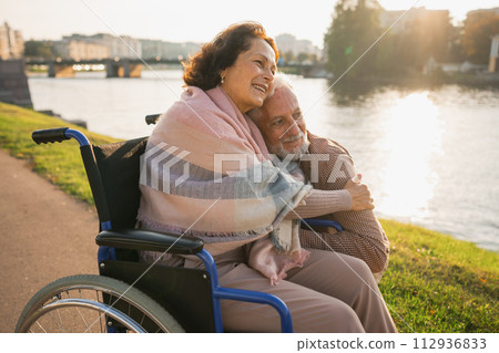 Senior woman in wheelchair walking with caregiver old man on road in park. Elderly family couple man supporting embracing woman in chair for people with disability outdoor. Rehabilitation 112936833