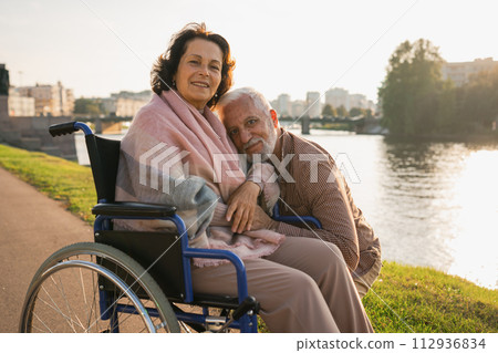 Senior woman in wheelchair walking with caregiver old man on road. Elderly family couple man supporting taking care of paralyzed woman in chair for people with disability outdoor. Rehabilitation 112936834