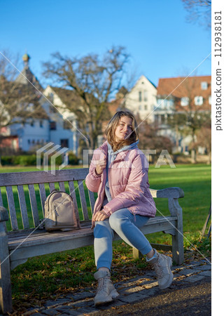 Winter Joy in Bitigheim-Bissingen: Beautiful Girl in Pink Jacket Sitting Amidst Half-Timbered Charm. beautiful girl in a pink winter jacket sitting on a bench in a park, set against the backdrop of 112938181