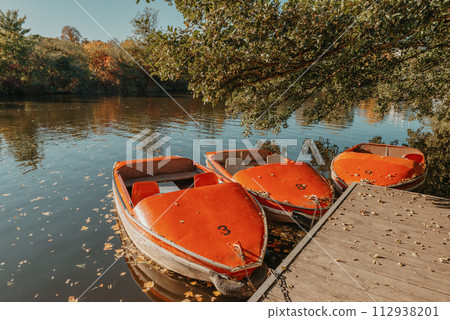 Several boats with oars are moored at the water's edge at the pier in the city park for water walks on the river, lake or pond. Sunset. Several boats with oars are moored at the water's edge at the pier in the city park for water walks on the river, lake or pond. Sunset. 112938201