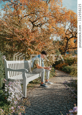 A Teenager Sits On A Bench In The Autumn Park Drinks Coffee From A Thermo Mug And Looks Into A Phone. Portrait Of Handsome Cheerful Guy Sitting On Bench Fresh Air Using Device Browsing Media Smm 112938225