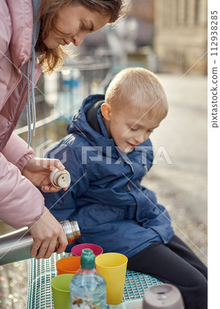 Family Picnic Delight: Cheerful 8-Year-Old Blond Boy in Blue Winter Jacket Sits on Bench While Mom Pours Tea from Thermos, Autumn or Winter. a joyful 8-year-old blond boy in a blue winter jacket Family Picnic Delight: Cheerful 8-Year-Old Blond Boy in Blue Winter Jacket Sits on Bench While Mom Pours Tea from Thermos, Autumn or Winter. a joyful 8-year-old blond boy in a blue winter jacket 112938285